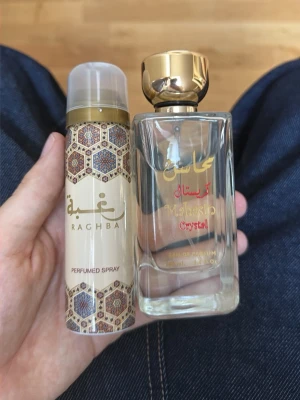 Raghba och Mahasin Crystal parfym - Två parfymer: en Raghba perfumed spray i beige flaska med orientaliskt mönster och en Mahasin Crystal Eau de Parfum i genomskinlig glasflaska med guldfärgad kork och text i guld och rött. Båda har en elegant och lyxig design.