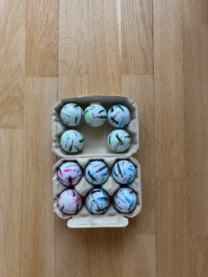 11 Splash golfbollar A klass - Säljer ett set med färgglada golfbollar från TaylorMade, förvarade i en äggkartong. Bollarna har olika färgade markeringar i blått, rosa och grönt samt svarta streck. Perfekt för dig som vill ha unika golfbollar på banan. Spelar som vanligt eller bara bättre med bollarna