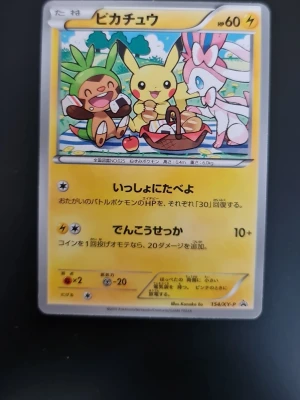 Pokemon Pikachu, chespin & Sylveon Picnic time - Unikt japanskt Pokémonkort med Pikachu, Chespin och Sylveon på picknickmotiv. Kortet har gul ram, färgglad framsida och klassisk blå baksida med Pokéball. Materialet är glansig kartong och texten är på japanska. Perfekt för samlare eller Pokémonfans.