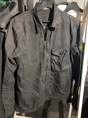 Svart Stone Island overshirt med dragkedja - Stilren svart overshirt med dragkedja framtill och en bröstficka med knapp. Klassisk krage och diskret patch på ärmen. Nypris ca 5000