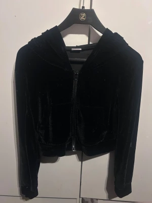 Svart velour hoodie med dragkedja - Säljer en svart croppad hoodie i mjuk velour med dragkedja framtill och huva. På ryggen står det 'Juicy' i glittriga stenar. Perfekt för dig som gillar en chill och trendig stil. Långa ärmar och fickor framtill.