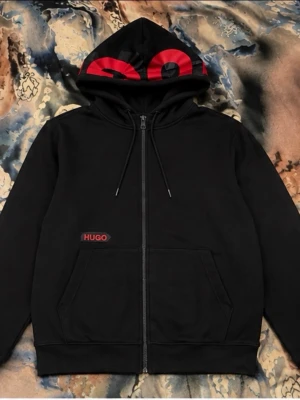 Svart hoodie med dragkedja från Hugo - Säljer en svart hoodie från Hugo med dragkedja och huva. Stort rött Hugo-tryck på huvan och mindre logga på bröstet. Praktiska fickor framtill och justerbar dragsko i huvan. Perfekt för en cool och avslappnad stil.