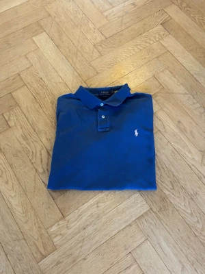 Blå pikétröja från Polo Ralph Lauren - Klassisk blå pikétröja från Polo Ralph Lauren med vit broderad logga på bröstet. 