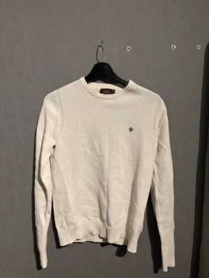 Vit stickad Morris tröja med broderad detalj - Klassisk vit stickad tröja med rund hals och diskret blå broderad logga på bröstet. Tröjan har ribbade muddar vid ärmslut och nederkant, samt en enkel och stilren design som passar till det mesta. Perfekt för lager på lager under kyliga dagar.