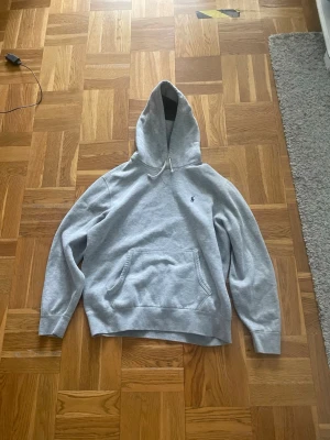 Grå hoodie från Polo Ralph Lauren - Snygg grå hoodie från Polo Ralph Lauren med klassisk broderad logga på bröstet. Hoodien har huva med dragsko och en stor magficka. Perfekt för en avslappnad och stilren look. Mjuk bomullsblandning som känns skön mot huden.