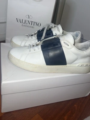 Valentino vita sneakers med svart detalj - Snygga vita sneakers från Valentino med en bred svart läderdetalj över sidan. Klassisk låg modell med vita snören och diskreta nitar på hälen. Tillverkade i slätt läder för en clean och exklusiv look. Det är en del slitage/hål på undersidan. Det går dock att laga. Även slitage på själva skorna förekommer. 