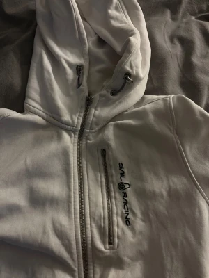 Vit hoodie från Sail Racing - Vit hoodie med dragkedja från Sail Racing. Har en bröstficka med dragkedja och broderad logga på vänster sida. Justerbar huva med snören och mjukt material som känns skönt mot huden. Perfekt för en avslappnad och sportig stil.