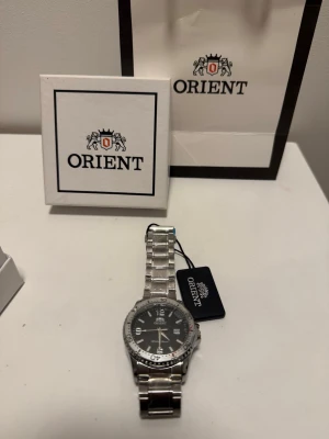 Orient silverfärgad klocka - Stilren klocka från Orient med svart urtavla och silverfärgat armband i metall. Klassisk rund form med tydliga siffror och datumvisning. Perfekt accessoar för dig som gillar en tidlös look.