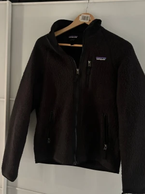 Svart fleecejacka från Patagonia - Mysig svart fleecejacka från Patagonia med hög krage och hel dragkedja framtill. Jackan har tre fickor med dragkedja och en klassisk, enkel design. Perfekt för kyliga dagar och enkel att matcha med olika outfits. Lite små nopprig men inget man tänker på vid användning och en defekt längs ner på en ärm men som går att vika upp så det inte märks!
