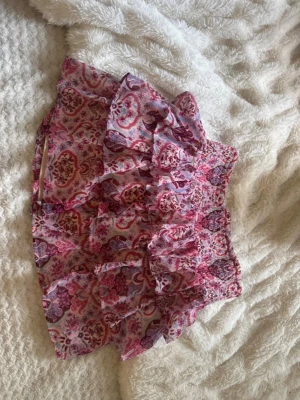 Rosa volangkjol med blommigt mönster - Söt kort volangkjol i tunnt material med blommigt mönster i rosa, lila och vitt. Kjolen har flera lager volanger och bred resår i midjan för en bekväm passform. Perfekt för sommarens alla tillfällen. 