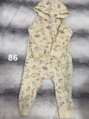 Gul blommig jumpsuit med huva - Jumpsuit i gult tyg med blommigt mönster i rosa och grönt. Modellen har huva, långa ärmar, muddar vid ärmar och benslut samt dragkedja längs sidan. Perfekt för en chill och bekväm look.🌸 Fläckar. Storlek 86