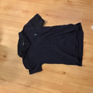 Mörkblå pikétröja från Polo Ralph Lauren - Klassisk mörkblå pikétröja från Polo Ralph Lauren med den ikoniska broderade loggan på bröstet. Tröjan har krage, korta ärmar och knappar vid halsen. Perfekt för en stilren och avslappnad look.