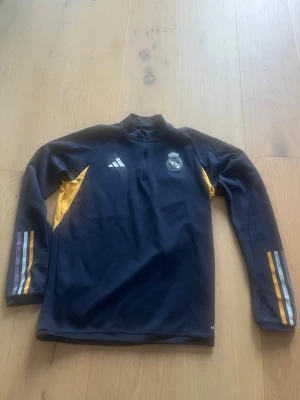 Real Madrid träningströja Adidas M - Mörkblå långärmad träningströja från Adidas med Real Madrid-logga på bröstet. Tröjan har gula detaljer på sidorna och randiga ärmar i gult, vitt och lila. Perfekt för fotbollsträning eller chill. Halv dragkedja framtill.Pris kan diskuteras.