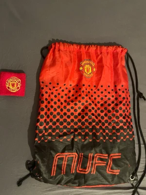 Manchester United gympåse röd/svart och svettband - Snygg gympåse i rött och svart med Manchester Uniteds logga och texten MUFC. Tillverkad i lätt syntetmaterial med dragskostängning och svarta snören. Perfekt för träning eller skola för dig som älskar fotboll och vill visa ditt lag, och ett svettband, 