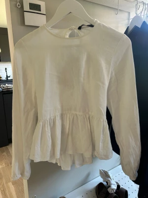 Vit blus med öppen rygg och volang - Supersöt vit blus i tunt, luftigt tyg med lång ärm och volang nedtill. Blusen har en snygg öppen rygg med knytband och cut-out detaljer. 
