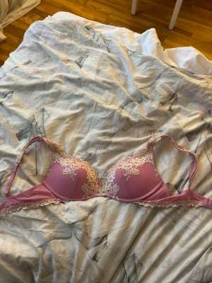 Rosa bh med spetsdetaljer - Söt rosa bh med vadderade kupor och fina spetsdetaljer i vitt framtill. Justerbara axelband och hakknäppning bak. Materialet är en mix av mesh och spets som ger en romantisk känsla.