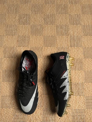 Svarta Nike fotbollsskor med dobbar - Säljer ett par svarta Nike fotbollsskor med vita detaljer och guldfärgade dobbar. Skorna har en låg profil, snörning och mönstrad sula för bra grepp på planen. Snygg design med grafiska inslag och texten 'NIKESKIN' på sidan.