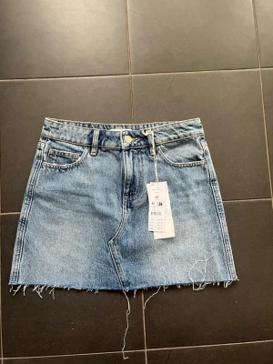 Kort jeanskjol i ljusblå denim - Säljer en kort jeanskjol i ljusblå denim med rå kant nertill och klassiska fem fickor. Kjolen har knapp och dragkedja framtill samt bälteshällor. Perfekt för dig som gillar en avslappnad och trendig stil. Helt ny och oanvänd! 