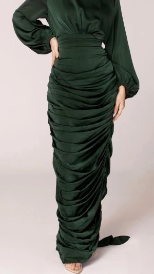 Mörkgrön långklänning i satin - Elegant mörkgrön långklänning i glansigt satinmaterial med markerad midja och draperad kjol. Klänningen har långa puffärmar, bred knyt i midjan och en snygg omlottkänsla baktill. Perfekt för dig som vill sticka ut med en lyxig och sofistikerad look. Klänningen är från Veiled collection och är storlek M.