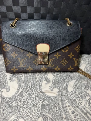  axelremsväska - Snygg handväska med klassiskt monogrammönster i brunt och guld. Väskan har svart lock i strukturerat läder, guldfärgad kedjerem och ett elegant metallspänne framtill. Perfekt accessoar för dig som gillar lyxig och tidlös design.