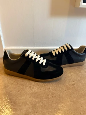 Maison Margiela Gats - Stilrena svarta sneakers med kontrasterande beige yttersula och vita skosnören. Skorna har en klassisk låg profil och detaljer i både mocka och slätt material. Perfekta för en avslappnad och trendig look.