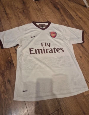 Arsenal bortatröja Nike vit - Snygg vit Arsenal bortatröja från Nike med vinröda detaljer och klubbmärke på bröstet. Tröjan har Fly Emirates-tryck på framsidan och är tillverkad i lätt, andningsbart material med NikeFIT-teknologi. Perfekt för fotboll eller som supporterplagg. Sitter som M jag är 181 