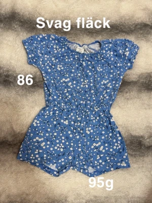 Blå blommig jumpsuit med kort ärm - Söt blå jumpsuit med vita små blommor och korta ärmar. Resår i midjan för skön passform och knytning bak i nacken. Perfekt för varma dagar och enkel att ta på. Lätt och luftig modell.🌸 Svag fläck. Storlek 86