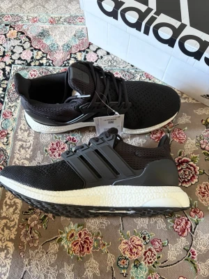 Adidas Ultraboost sneakers - Svarta Adidas Ultraboost sneakers med stickad ovandel och vita Boost-sulor. Skorna har klassiska tre ränder på sidan, svart hälkappa och snörning. Perfekta för dig som gillar stilrena och bekväma sneakers med sportig känsla.