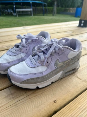 Nike Air Max lila och vita sneakers - Nike Air Max sneakers i lila och vitt med silverdetaljer. Skorna har en klassisk låg siluett, synlig Air Max-dämpning i sulan och är tillverkade i en mix av mocka, textil och syntet. Perfekta för dig som gillar en sportig och trendig look.