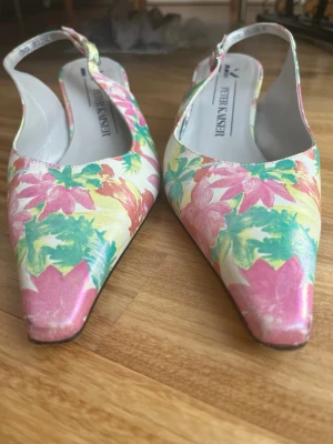 Blommiga pumps med låg klack - Säljer ett par färgglada pumps från Peter Kaiser med blommigt mönster i rosa, grönt och gult. Skorna har spetsig tå, låg klack och öppen häl med justerbar rem. Perfekta för dig som vill sticka ut med en unik och somrig look.