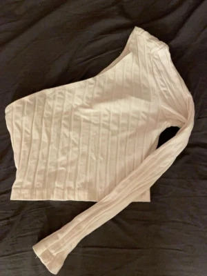 Beige oneshoulder topp med ribbad struktur - Trendig beige oneshoulder topp med ribbad struktur och lång ärm. Snygg croppad passform som ger en modern look. Perfekt att styla med höga jeans eller kjol för en cool outfit.