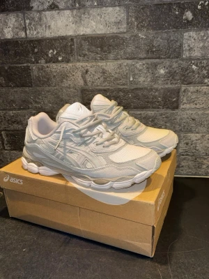 Vita Asics GEL-NYC  - Säljer ett par fräscha vita Asics sneakers med cool chunky sula. Skorna har en blandning av mesh och syntetläder för både stil och komfort. Klassisk Asics-logga på sidan och vita skosnören. Perfekta för dig som gillar sportig och clean look.