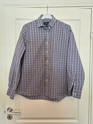 Rutig skjorta från Hampton Republic - Säljer en klassisk rutig skjorta från Hampton Republic i regular fit. Skjortan har långa ärmar, knappar framtill och ett mönster i blått, vitt och brun Perfekt för dig som gillar en tidlös och stilren look.