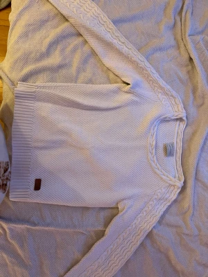 Vit stickad tröja med flätade ärmar - Vit stickad tröja med rund hals och flätmönster längs ärmarna. Tröjan har ribbade muddar och en liten brun patch nertill. Materialet ser ut att vara mjukt och tjockt, perfekt för kyligare dagar.