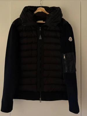 Svart cardigan hybrid jacket  - Snygg svart cardiganjacka från Moncler med vadderad framsida och stickade ärmar. Jackan har huva, hel dragkedja och en ficka med dragkedja på ärmen. Moncler-logga på vänster ärm. Perfekt för kyliga dagar och riktigt trendig look, Med nfc Scan som tar dig till hemsidan.