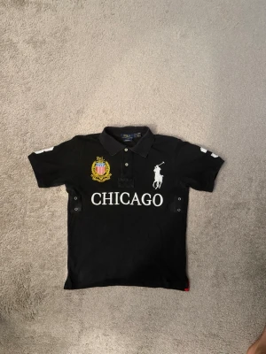 Svart piké med CHICAGO-tryck - Snygg svart pikétröja från Ralph Lauren med stort vitt CHICAGO-tryck på framsidan. Säljer den för att den är för liten för mig. Letar också efter byten som är storlek M