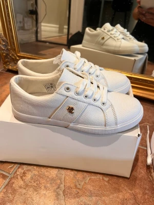 Vita Ralph Lauren sneakers  - Snygga vita sneakers i tyg med vita snören och gummisula. Diskret guldfärgad detalj på sidan och klassisk låg profil som passar till det mesta. Bekväma och fina. Endast använda en kort stund. Säljes pga att jag köpte fel storlek. Nypris ca 1300kr