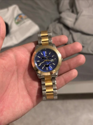 Blå klocka med gulddetaljer - Snygg klocka med blå urtavla och silver- samt guldfärgad länk. Klockan har markerade index och visare, samt en robust boett i metall. Perfekt accessoar för dig som vill sticka ut med stilrena detaljer. Fråga för mer bilder 