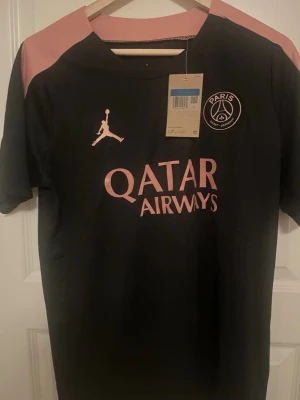 Svart PSG matchtröja Jordan M - Snygg svart PSG matchtröja från Jordan med rosa detaljer och logga på bröstet. Tröjan har korta ärmar, rund hals och tryck med Qatar Airways på framsidan. Perfekt för fotboll eller streetwear. Tillverkad i lätt och ventilerande material.