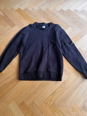 Svart långärmad tröja C.P. Company - Svart långärmad tröja från C.P. Company i mjuk bomull. Klassisk rund halsringning och en unik detalj med ficka och glaslins på ena ärmen. Perfekt för dig som gillar stilrena plagg med en twist.