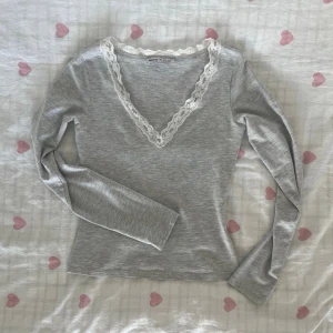 Grå långärmad topp med spets - Söt grå långärmad topp med v-ringning och vit spetskant runt halsen. Toppen har en slim passform och är tillverkad i mjukt bomullsmaterial. Perfekt för dig som gillar en enkel men ändå romantisk stil.