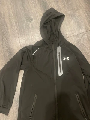 Svart vindjacka från Under Armour - Svart vindjacka från Under Armour med huva, hel dragkedja och flera fickor med dragkedja. Jackan har en vit logga på bröstet och en grå detalj vid bröstfickan. Perfekt för blåsiga dagar och sportiga outfits.