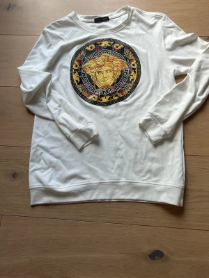 Vit sweatshirt med Medusa-tryck - Snygg vit sweatshirt med ett stort, färgstarkt Medusa-tryck i gult, svart och rött på bröstet. Tröjan har rund hals, långa ärmar och ribbade muddar. Perfekt för dig som vill sticka ut med ett ikoniskt motiv.