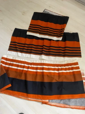 Randiga gardinkappor i orange och svart - Säljer tre gardinkappor med breda och smala ränder i orange, svart och vitt. Tyget är bomull och har en klassisk retrostil. Perfekt för att ge köket eller vardagsrummet en färgklick och unik känsla.