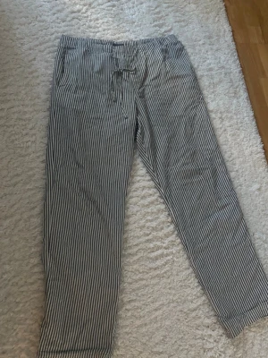 Randiga pyjamasbyxor  - Säljer ett par klassiska pyjamasbyxor med tunna vita och mörkblå ränder. Byxorna har rak passform, resår i midjan och justerbar dragsko för extra komfort. Perfekta för slappa dagar hemma eller mysiga kvällar.