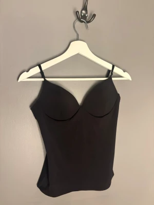 Svart body med tunna axelband - Snygg svart body med tunna justerbara axelband och formpressade kupor. Materialet är slätt och stretchigt för en bekväm passform. Perfekt att bära under kläder eller som ett statement-plagg. Enkel och stilren design som passar till mycket.