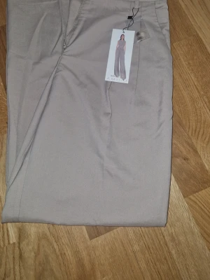 Beige vida kostymbyxor - Snygga beige kostymbyxor med vida ben och hög midja. Byxorna har klassisk dragkedja och knapp framtill. Perfekta för en stilren och trendig look. Materialet ser ut att vara mjukt och följsamt, vilket ger en skön känsla.nya