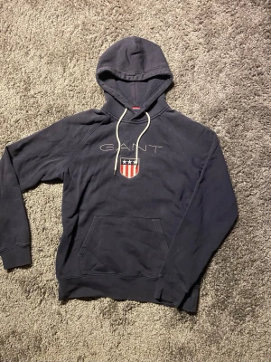 Mörkblå hoodie från GANT med logga - Mörkblå hoodie från GANT med vit dragsko i huvan och stor ficka framtill. På bröstet finns GANT-logga och ett broderat sköldmärke i rött, vitt och blått. Hoodien har ribbade muddar vid ärmslut och nederkant. Perfekt för en avslappnad och stilren look.