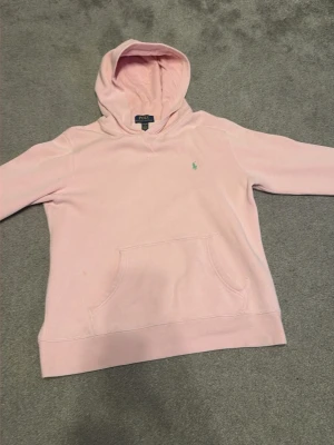 Hoodie från Polo Ralph Lauren - Säljer en ljusrosa hoodie från Polo Ralph Lauren med klassisk huva och känguruficka framtill. Tröjan har det ikoniska lilla broderade logotypen på bröstet och är gjord i mjukt bomullsmaterial. Perfekt för en avslappnad och stilren look.