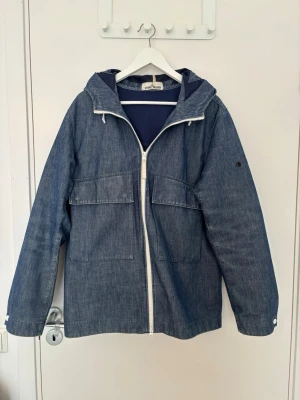 Stone Island jacka  - Snygg blå vindjacka i denim-look med vit dragkedja och stora fickor framtill. Jackan har huva, långa ärmar och knappar vid ärmslut. Perfekt för blåsiga dagar och enkel att matcha med olika outfits.